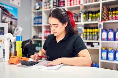 Preview: young latin woman behind counter working, using calculator and writing in small auto parts store