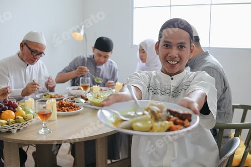 Preview: Young Asian Muslim Man Showing His Food to Camera Against Blurry Family Eating Together Background