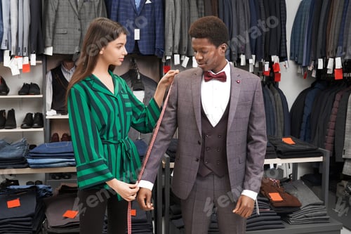 Preview: Man fitting grey suit, assistant measuring sleeve with tape