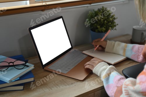 Preview: Young woman making notes on notebook and using computer laptop at home.