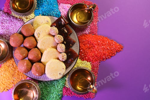Preview: Diwali celebration. Tasty Indian sweets, diya lamps and colorful rangoli on violet table, flat lay