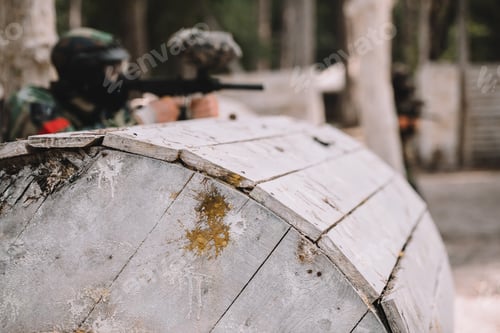 Preview: selective focus of paintball splashes on wooden barrel and paintball player shooting behind outdoors