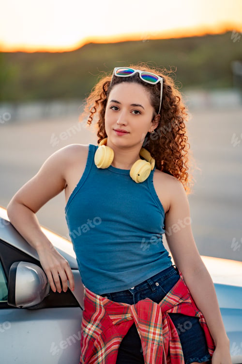 Preview: Young woman with headphones relaxing by a car at sunset.