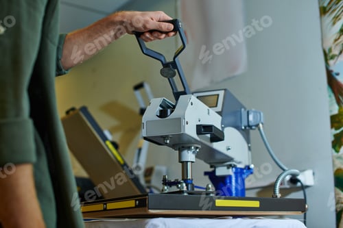 Preview: Cropped view of young craftsman using screen printing machine on clothes while working in blurred