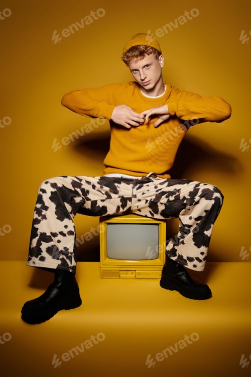 Preview: young man with red hair models his trendy winter outfit and sitting on vintage tv