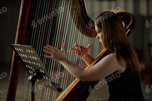 Preview: Young Woman Strumming Harp Strings Playing Classical Music in Dim Light Indoors