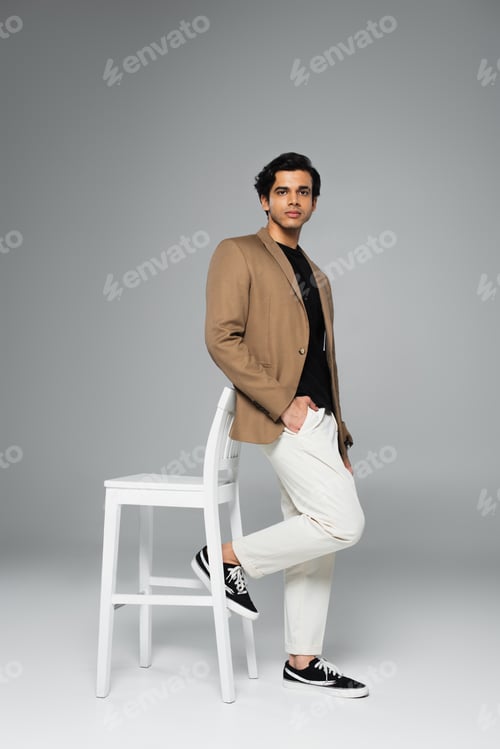 Preview: full length of young man in blazer posing with hand in pocket near white chair on grey