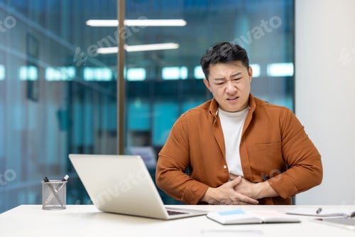 Preview: Asian businessman experiencing stomach discomfort at office desk while working on laptop