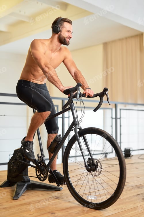 Preview: Cheerful athletic man riding stationery bicycle and listening to music