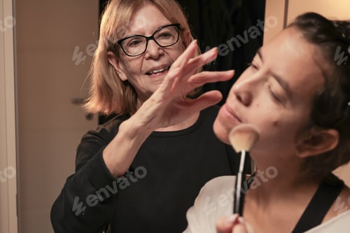 Preview: Woman applying makeup with brush assisted by elderly woman smiling. Mother's Day