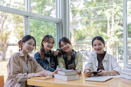 Preview: University students sitting together at table with book and laptop. Happy young people doing group