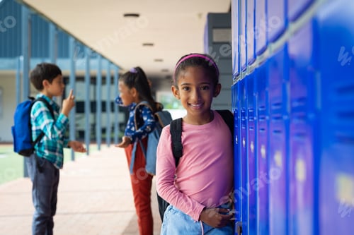 Preview: Biracial girl with a pink backpack smiles near school blue lockers, her dark hair tied back