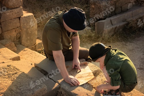 Preview: Two archeologists, child and adult, in khaki clothes sitting on ruins and studying an ancient map.