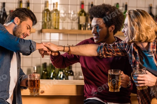 Preview: smiling young multiethnic men holding glasses of beer and stacking hands in bar