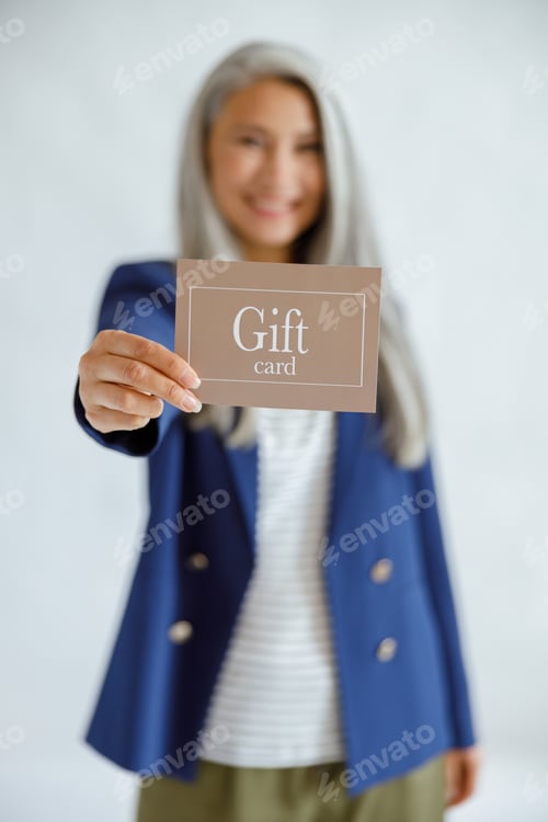 Preview: Positive middle aged Asian woman on light grey background, focus on hand with gift card