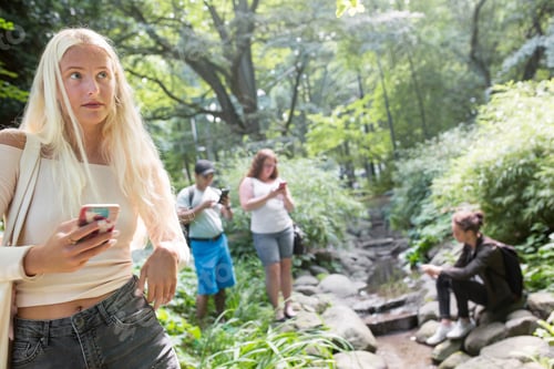 Preview: Woman checking her phone in a lush green park with others enjoying the surroundings.