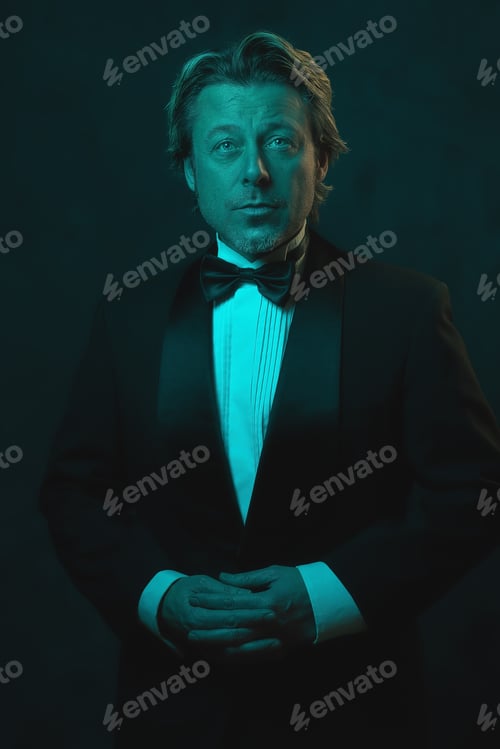 Preview: Blonde Man Wearing Tuxedo And Bow Tie. Lit By Blue And Orange Light Against Dark Wall.