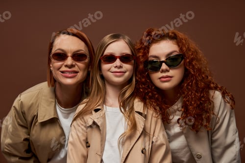 Preview: portrait of happy three generations of women looking at camera in sunglasses on brown backdrop