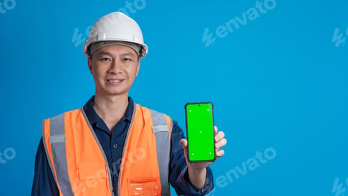 Preview: Confident young engineer wearing orange and a safety helmet stands holding a laptop, phone