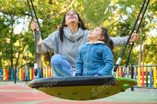 Preview: Happy Latin mother playing with her son looking up on a swing in a park.
