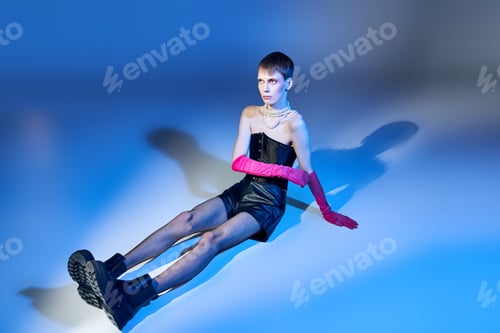 Preview: model in corset and boots sitting on blue backdrop, nonbinary person in pink gloves, queer fashion