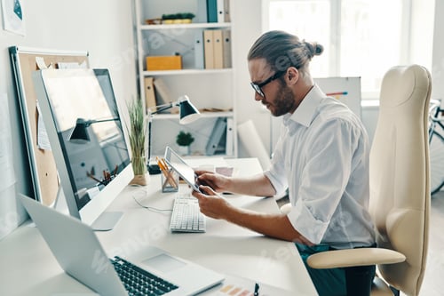 Preview: Full concentration. Good looking young man in shirt using digital tablet while sitting in the office