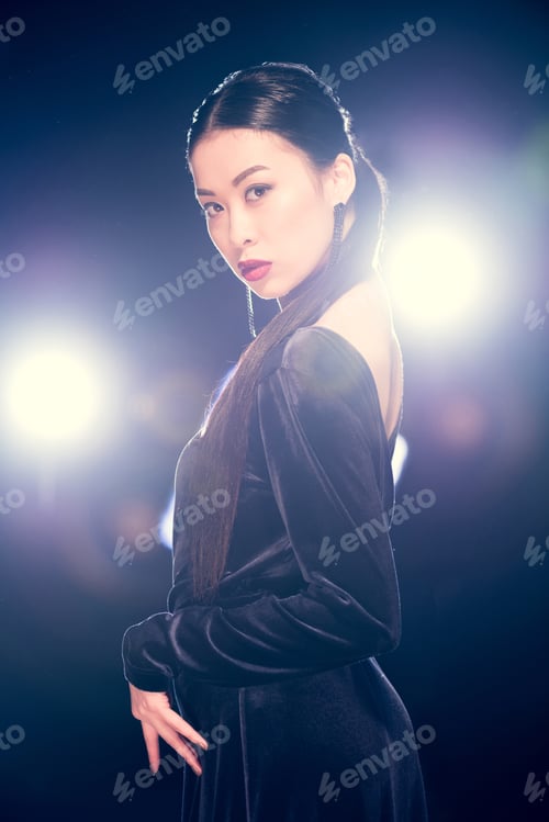Preview: seductive asian woman posing in black evening dress and looking at camera at event with backlit