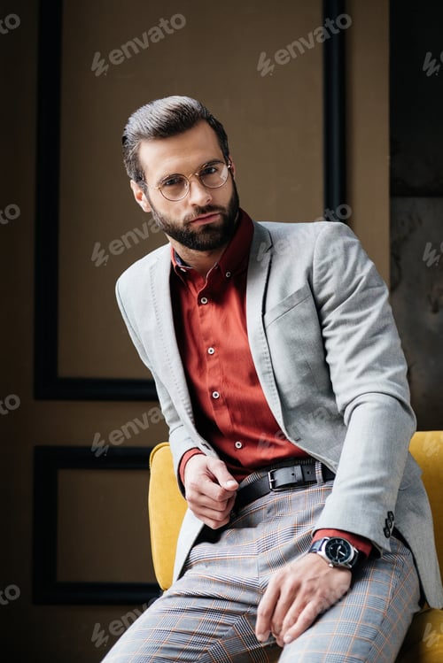 Preview: handsome bearded man in glasses and stylish suit sitting on couch