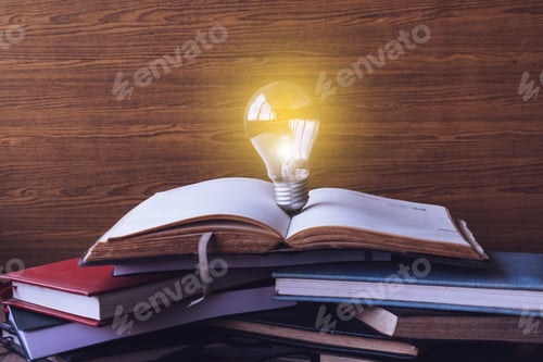 Preview: Open book with light bulb and hardback books on wood wall background. Selective focus