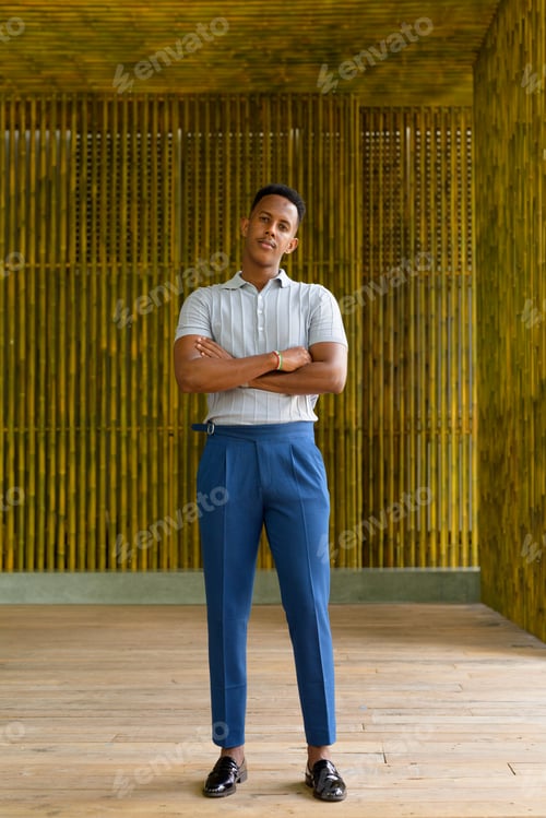 Preview: Full length shot of young African businessman smiling outdoors against bamboo wall with arms crossed