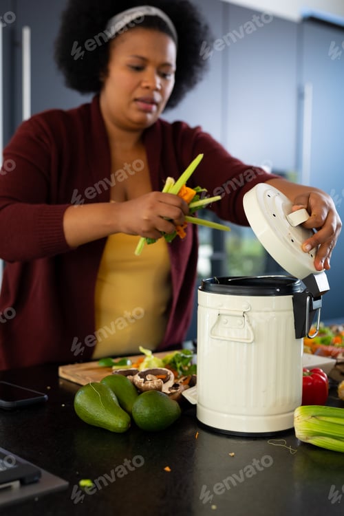 Preview: Happy plus size african american woman preparing dinner, composting vegetable waste in kitchen