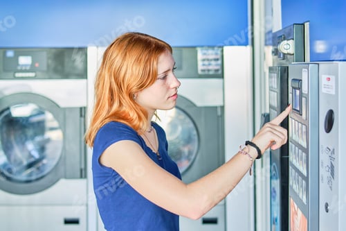 Preview: Young redhead adjusting the buttons on an automatic washing machine in a public laundromat