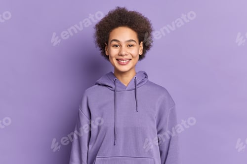 Preview: Happy ethnic teenager with Afro hair smiles positively wears purple hoodie being in good mood poses
