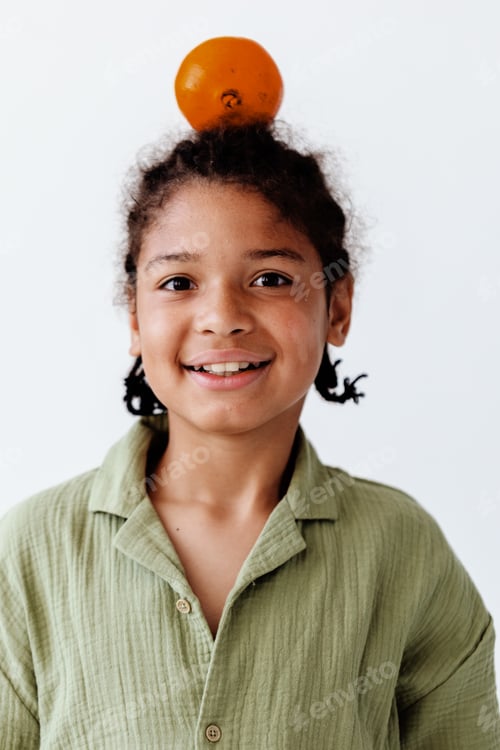 Visualização: retrato criativo e caprichoso de um jovem garoto afro-americano equilibrando uma laranja na cabeça