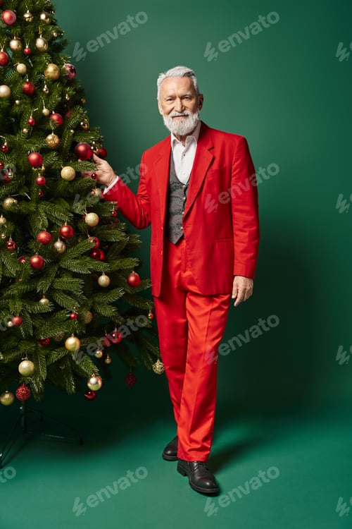 Preview: stylish handsome Santa Claus posing near Christmas tree touching decorations, winter concept