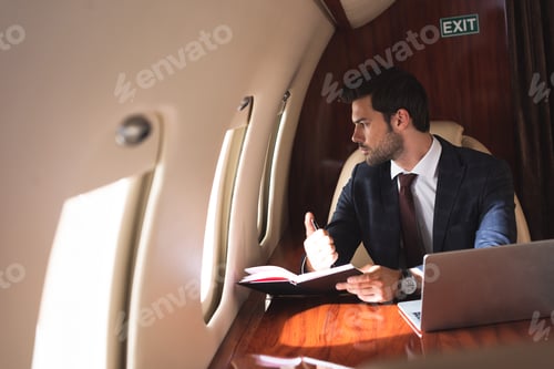 Preview: young businessman working with notebook and laptop in plane during business trip