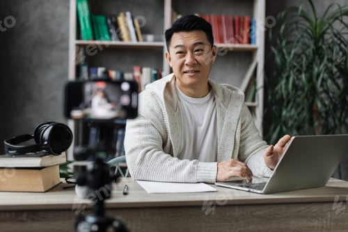 Preview: Attractive asian male working on laptop and recording video on digital smartphone