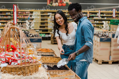 Preview: happy african american man holding metal scoop while taking raisins near asian woman in store