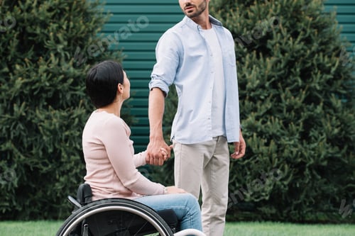 Preview: cropped view of young man holding hands with disabled girlfriend while walking in park together