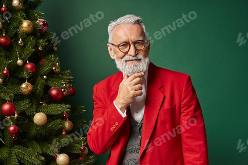 Preview: jolly Santa in glasses with beard in classy suit posing near tree with hand on chin, winter concept