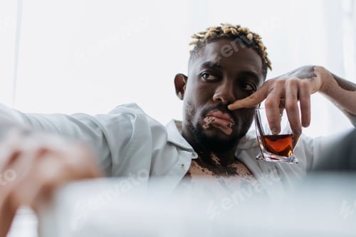 Preview: Young african american man with vitiligo in white shirt holding whiskey in blurred bathtub