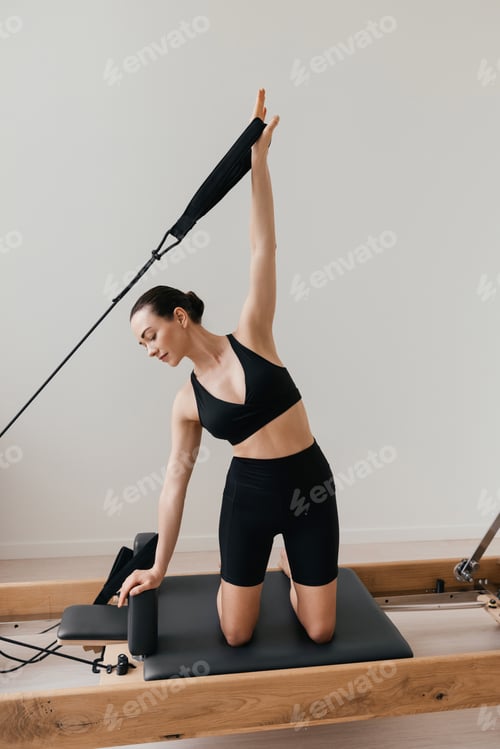 Preview: Slim brunette woman in black tight sportswear working out on Pilates reformer machine