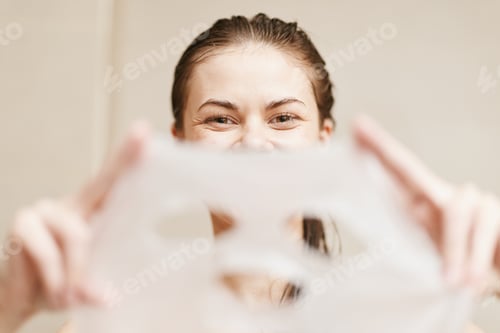 Preview: Joyful young woman holding a face mask, smiling playfully in a light-colored bathroom. Skincare and
