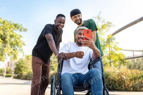 Preview: Young African American man in a wheelchair using phone together with male friends outdoors.