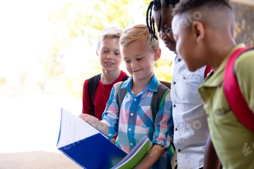 Preview: Caucasian elementary schoolboy showing books to multiracial male classmates at school