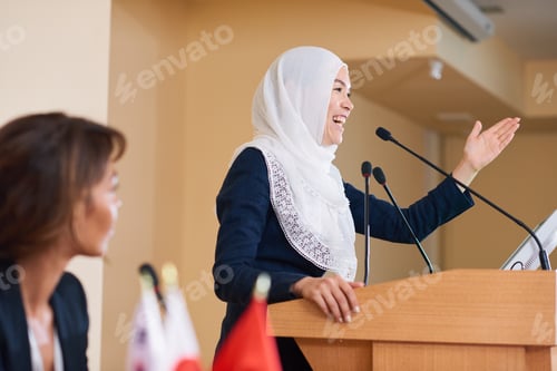 Preview: Happy young female speaker in hijab laughing while standing by tribune