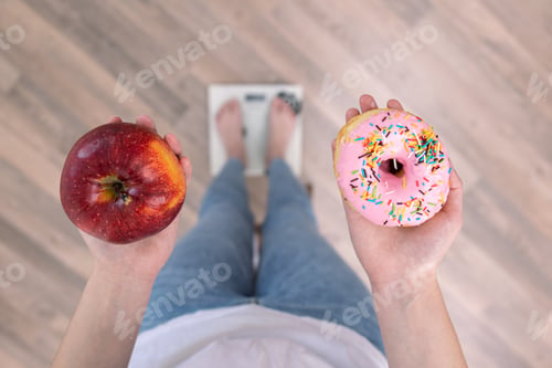 Preview: A woman stands on the scales, holds a donut and an apple in her hands, top view.