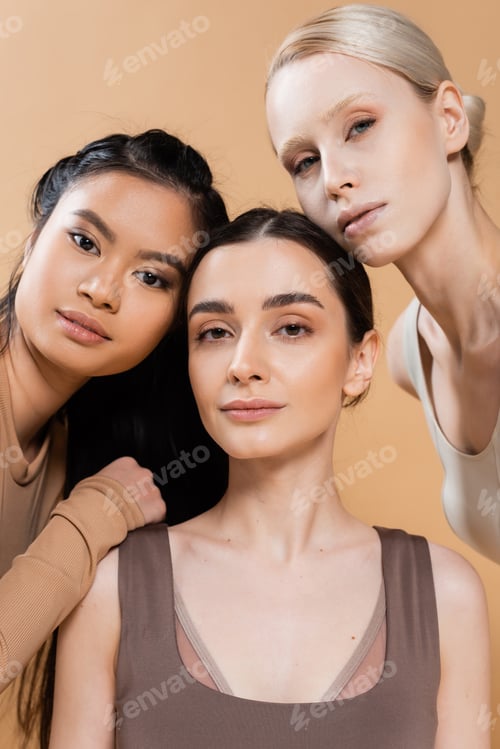Preview: young and pretty multiethnic women in underwear looking at camera isolated on beige