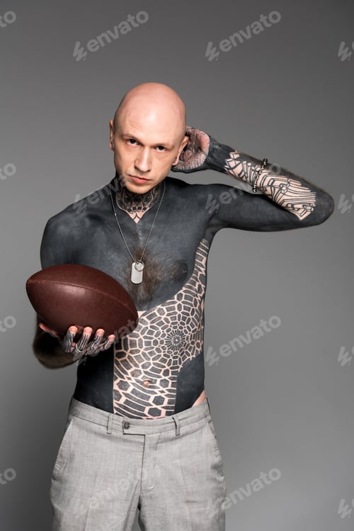 Preview: bald shirtless tattooed man holding rugby ball and looking at camera isolated on grey