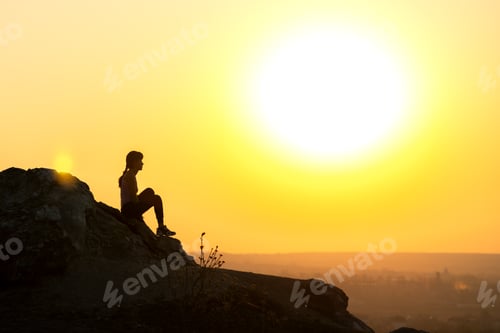 Preview: Silhouette of a woman hiker sitting alone on big stone at sunset in mountains
